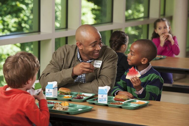 Schools Work to Make Cafeteria Lunches Healthier Children eat a balanced lunch together at tables in a bright school cafeteria.